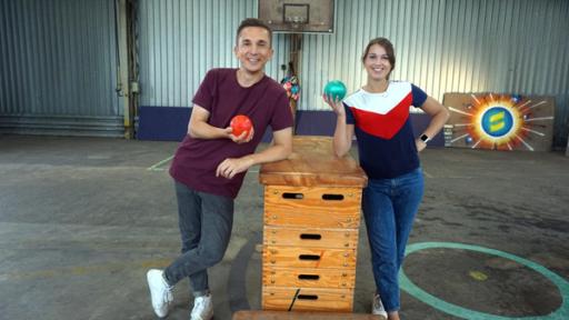 Die Sportmacher: Die Moderatoren Eric Mayer und Laura Knöll lehnen sich an einen Sprungkasten, der in ihrer Mitte steht. Beide halten Wasserbomben in der Hand. 