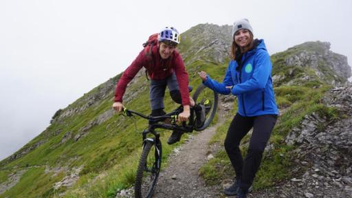 Tom Öhler (links) springt mit seinem Bike und Laura (rechts) steht nebendran. Die beiden stehen auf einem Berg.