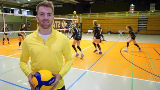 Die Sportmacher: Stefan steht in einer Sporthalle, im Hintergrund läuft ein Volleyballspiel. Er selbst hat einen Volleyball in der Hand. 