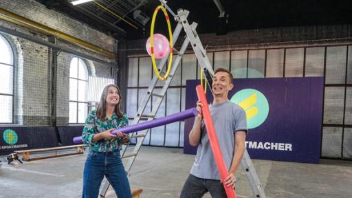 Die Sportmacher: Laura und Gastmoderator Eric Mayer in der Sportmacher-Halle. Beide halten eine Schwimmnudel in der Hand. Im Hintergrund steht eine große Leiter und das Sportmacher-Logo.