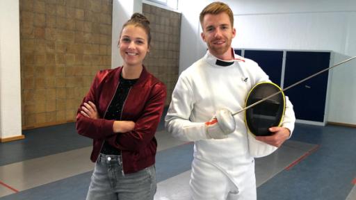 Die Sportmacher: Die Moderatoren Laura und Stefan stehen in der Turnhalle. Stefan trägt einen Fechtanzug und hält ein Florett in der Hand. Unter dem Arm trägt er eine Schutzhaube. Laura steht lachend mit verschränkten Armen neben ihm. 