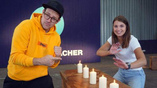 Die Sportmacher: Die Moderatoren Ben und Laura stehen neben einer Reihe brennender Kerzen und haben die Hände in Kampfkunst-Art erhoben. 