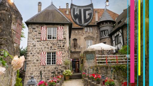 Ein Steinhaus mit roten Fensterläden, einem Turm, Blumen und einem großen Schild mit den Buchstaben WG. 