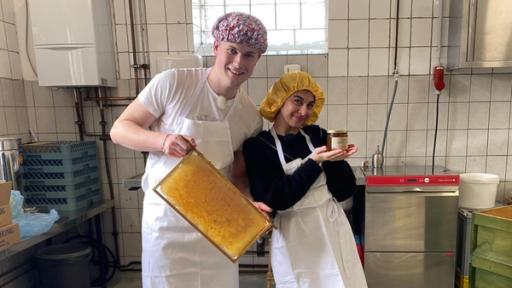 Die WG - Das große Wiedersehen: Mark und Ella aus der Jubiläums-WG stehen in einer weiß gekachelten Industrieküche. Beide tragen aus hygienischen Gründen weiße Schürzen und eine Haube über den Haaren. Ella präsentiert in ihren Händen ein Glas Honig. Mark hält einen Holzrahmen mit Bienenwaben in die Kamera. Beide lächeln.