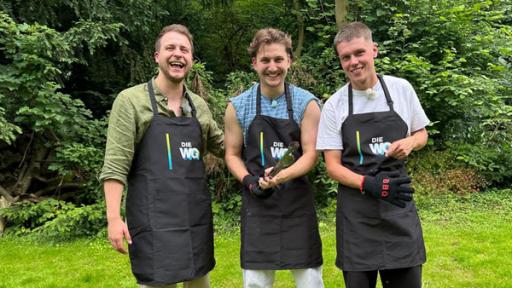 Die WG - Das große Wiedersehen: Jonas, Robin und Jeppe aus der Jubiläums-WG stehen in einem Garten mit Bäumen auf einer Wiese. Sie tragen alle schwarze Grillschürzen mit dem WG-Logo darauf. Robin, in der Mitte der Gruppe, hält eine Grillzange in den Händen. Jeppe, auf der rechten Seite im Bild, zieht Grillhandschuhe an. Alle lachen in die Kamera.