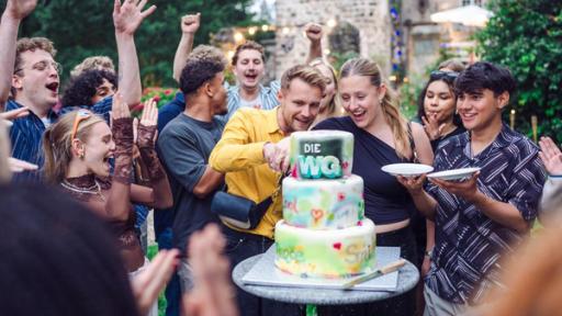 Die WG - Das große Wiedersehen: Im Mittelpunkt des Fotos steht eine dreistöckige, bunte Torte mit WG Logo. Aaron und Hanna aus der Jubiläums-WG stehen hinter der Torte und schneiden sie gerade an. Dominik steht rechts neben Hanna und hält erwartungsvoll zwei Kuchenteller bereit. Viele andere WGlerinnen und WGler stehen um die Gruppe herum, klatschen und jubeln und freuen sich offenbar auf die Torte. Im Hintergrund ist die Burg zu sehen, in der die Jubiläums-WG stattgefunden hat.