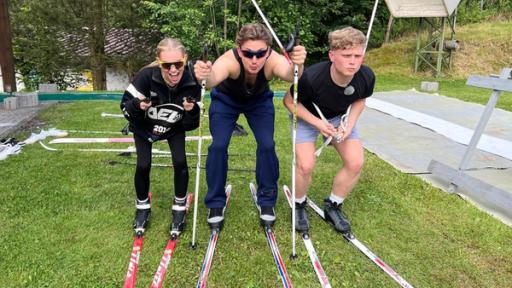 Die WG - Das große Wiedersehen: lexa, Robin und Julius stehen nebeneinander auf einer grünen Wiese und tragen Langlaufskier und Stöcke. Sie tun so, als würden sie mit Skiern gleich losfahren wollen. Alexa und Julius haben sich die Stöcke dafür unter die Arme geklemmt, Robin hat beide Stöcke in den Boden gesteckt, um sich vermeintlich abzustoßen. Alexa und Robin tragen Sonnenbrillen und lange Trainingshosen, Julius steht scheinbar konzentriert mit kurzer Hose und T-Shirt in Startposition.