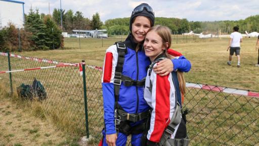 Toni und Alexa stehen in Fallschirmoutfit auf einem Flughafen