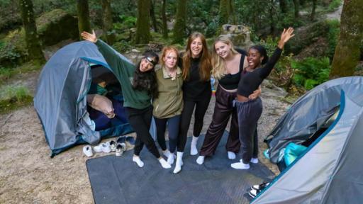 Fünf Mädchen stehen vor zwei Iglu-Zelten in einem Wald. Sie tragen Jogginghosen. Die Zelte sind offen, es liegen Schlafsäcke und Luftmatratzen darin. Vor den Zelten liegen die Turnschuhe der Mädchen. Die Fünf haben die Arme umeinander gelegt und lachen in die Kamera.