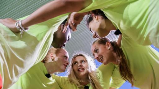 Fünf Mädchen in neongelben T-Shirts stecken als Team im Kreis die Köpfe zusammen und haben sich die Arme gegenseitig über die Schultern gelegt. Der Fotograf steht unterhalb von Ihnen und blickt nach oben in die eifrigen Gesichter. Die fünf Mädchen besprechen offenbar eine Spiel-Taktik. Die Mädchen wirken etwas zerzaust, aber fröhlich.