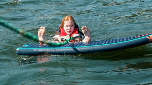 Mitten im Waser liegt ein Waveboard. Dahinter ist der Kopf eine rothaarigen Mädchens zu sehen, sie hält in der Hand eine grüne Zugleine. Die Füße streckt sie nach vorne in die Kamera, die Fersen liegen an der Kante des Boards auf.