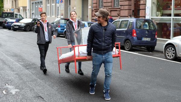 Auf der Suche nach einem Schlafplatz: Tupac, Sarah und Moderator Tommy Scheel tragen das rote Bett durch Kölns Straßen. | Rechte: ZDF/Annalena Renneisen