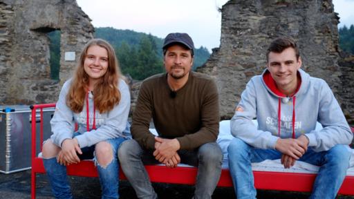 Tommy sitzt mit zwei Jugendlichen vom Draußen schlafen-Team auf einem roten Metallbett. Alle schauen erwartungsvoll in die Kamera.