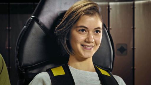 Groß im Bild ist Astronautin Emma glücklich lächelnd, angeschnallt im Sitz des Beschleunigungs-Simulators.