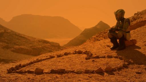 Eine Astronautin sitzt rechts im Bild neben einem großen Herz aus Steinen, links im Bild. Vom Hügel blickt man in die bergige, rötliche, sonnenbeschienene Marslandschaft.