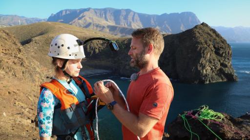 Wildniscoach Tobi zieht Tania die Klettersicherung an und zieht sie fest. Tania trägt einen Helm mit Kamera. Im Hintergrund ist eine Felsbucht und das Meer zu sehen.