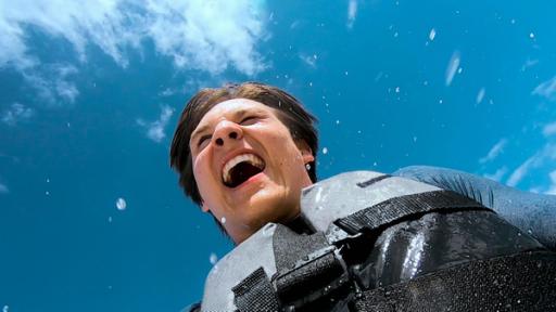 Ben hat eine Schwimmweste an und steht lachend im spritzenden Meerwasser.