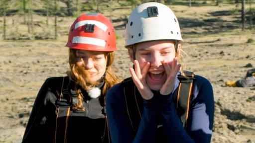 Toni und Tania stehen nebeneinander und haben Helme auf. Tania hält sich ihre Hände zu einem Trichter geformt vor den Mund und feuert die anderen an.