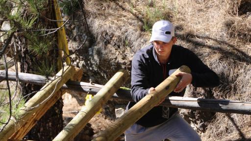 Bruno steht mit Schirmkappe am Gerüst einer Holzplattform und befestigt eine Holzstrebe.