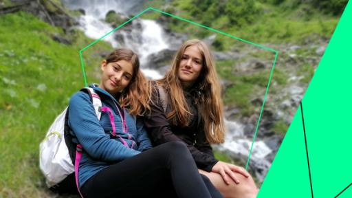 Elena (l.) und Johanna (r.) sitzen auf einem, am Boden liegenden, Baumstamm. Im Hintergrund: der Wasserfall Jungfernsprung.