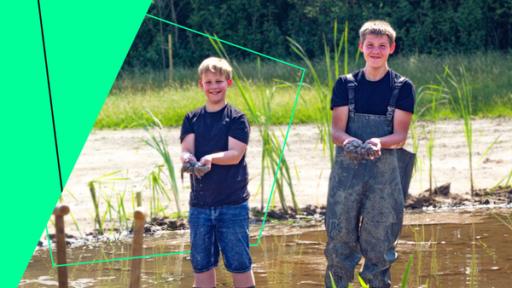 Lukas (l.) und Nico (r.) stehen mit Gummistiefeln und Anglerhose in einem Regenrückhaltebecken. Sie halten ihre mit Schlamm gefüllten Hände in die Kamera.