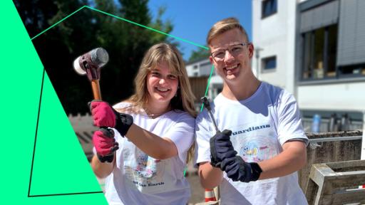 Annika (l.) und Lukas (r.) stehen auf einem Schulhof und halten Werkzeug in den Händen. Vor Ihnen liegt eine Palette und darauf Pflastersteine. Links im Bild steht ein gelber Bagger.