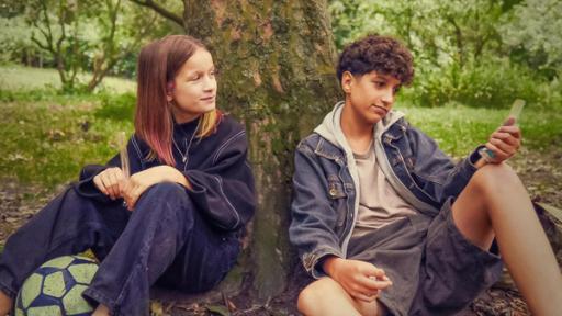 Liv und Pit sitzen im Grünen unter einem Baum. Beide haben ein Eis in einer schmalen durchsichtigen Tüte in der Hand. Vor Liv liegt ein Fußball. Liv blickt lächelnd und ein bisschen erwartungsvoll zu Pit, der leicht lächelnd nach vorne auf sein Eis schaut.