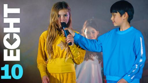 Im Bühnenlicht und von leichtem Nebel umgeben, stehen Celina (Lia Kahtin, l.) und Noah (Hauke Tài Hoàng) nebeneinander. Celina spricht in ein Mikrofon das Noah ihr vor den Mund hält. Im Hintergrund sieht man ein selbst gestaltetes Plakat und Leyla (Aleyna Kuş).