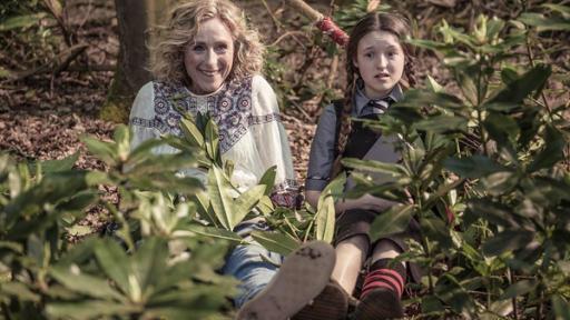 Mildred und ihre Mutter sitzen auf dem Waldboden. Julie lächelt und Mildred schaut verwirrt.