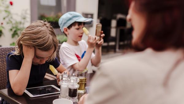 Zwei Kinder und eine Mitter sitzen an Tisch. Das Mädchen schaut auf ein Tablet, der Junge auf ein Smartphone.