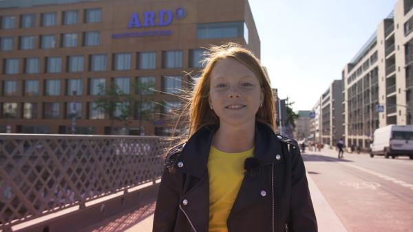 Celia steht vor einem Gebäude mit einem ARD-Logo darauf und lächelt in die Kamera.