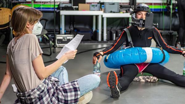 Rechts im Bild sieht man die Kikaninchen-Darstellerin Naemi. Sie trägt einen Motion-Capture-Anzug und sitzt auf dem Boden. Links neben ihr sitzt eine Redakteurin mit einem Zettel in der Hand.