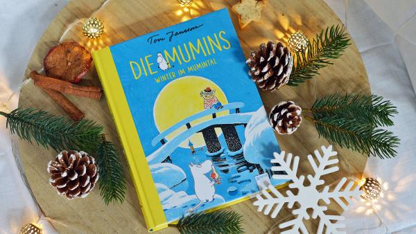 Das Buch "Die Mumins- Winter im Mumintal" von Tove Jansson liegt auf einem Holzbrett. Es ist blau mit gelber Schrift und der nilpferdartige Mumintroll ist darauf zu sehen. Um das Buch herum liegen Tannenzweige und Zapfen, sowie eine Lichterkette. Eine große gebastelte Schneeflocke liegt rechts unten daneben.