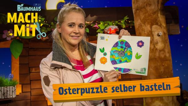 Eine blonde Frau (Singa) mit einer beigen Jacke und einem pink-weiß-gestreiften Pullover hält in der Hand die ausgemalte Bastelvorlage von einem Osterpuzzle.