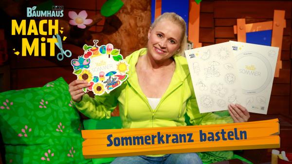 Eine blonde Frau mit einer grünen Jacke hält in der rechten Hand einen bunten Kranz aus Papier und in der linken eine Papiervorlage mit verschiedenen Ausmalmotiven darauf. 