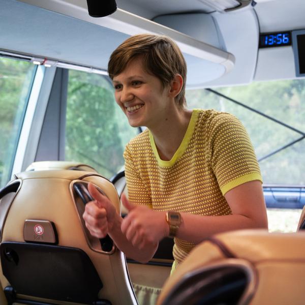 Eine Frau mit kurzen dunkelblonden Haaren steht zwischen den Sitzen eines Busses und zeigt erhobene Daumen.