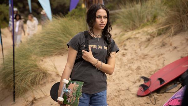 Am Strand: Summer (Sky Katz) steht mit ihrem Skateboard unter dem rechten Arm am Strand. Sie blickt skeptisch nach vorne. Neben ihr im Sand liegen Surfbretter und im Hintergrund sind Sand, Gräser und Menschen zu sehen.