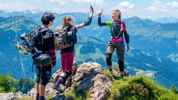 Drei Kinder stehen auf einem Berggimpfel. Zwei von ihnen geben sich eine High Five.