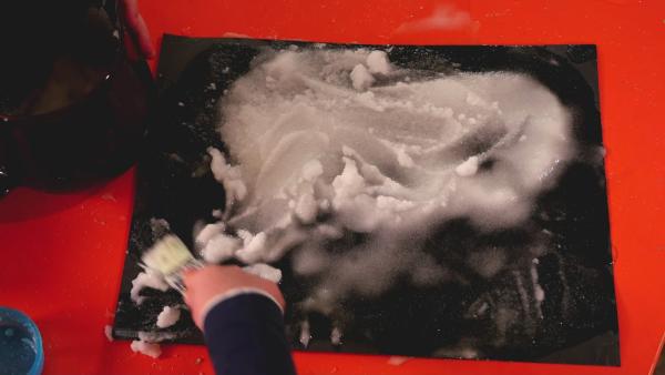 Auf einem schwarzen Tonpapier wird ein Salz-Wasser-Gemisch mit dem Pinsel aufgetragen. So entsteht ein Bild aus Salz. | Rechte: KiKA