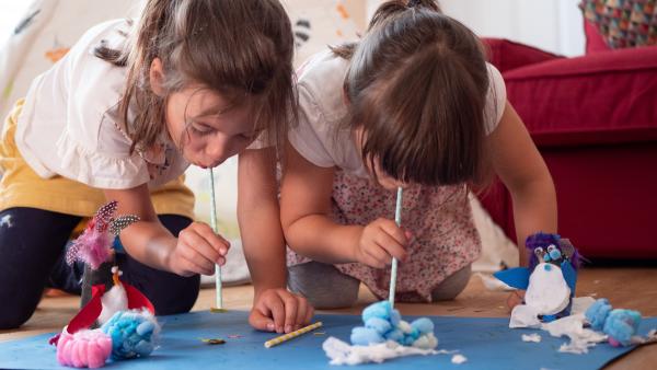 Zwei Mädchen mit Trinkhalmen versuchen Fische aus Papier in selbstgebastelte Pinguine aus Klopapierrollen zu heben.