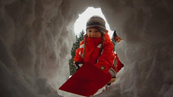 Ein Junge gräbt mit einer Schneeschippe ein Loch in einen Schneehaufen. | Rechte: KiKA