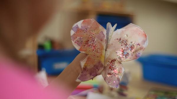 Ein mit rosa und lila Papierschnipseln beklebter Steckschmetterling von ENE MENE BU wird hochgehalten. Der Schmetterling ist mit viel Glitzer geschmückt. | Rechte: KiKA