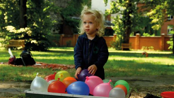 Hektor steht vor eine Kiste. In der Kiste sind viele bunte und mit Wasser gefüllte Luftballons. | Rechte: KiKA