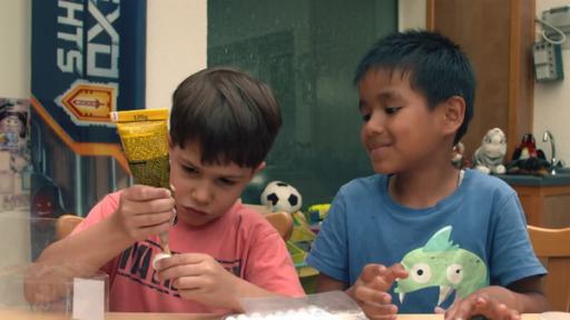 Zwei Kinder sitzen an einem Tisch und vor ihnen liegen Flaschendeckel und andere Bastelmaterialien.