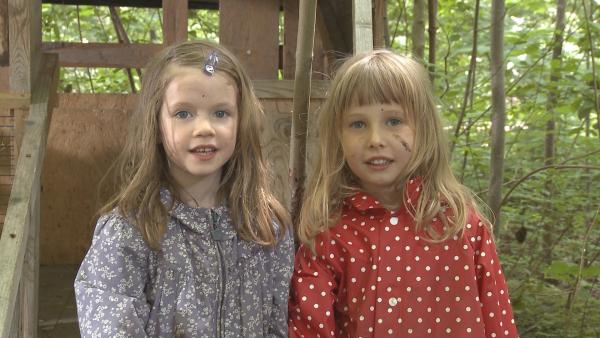 Zwei Mädchen stehen draußen vor einem Holzhaus. Sie sind im Wald und schmutzig im Gesicht.