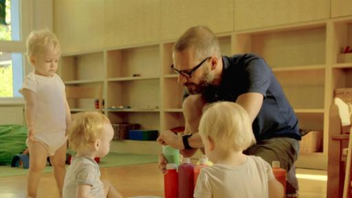 Drei Kleinkinder sitzen vor einem großen Blatt auf dem Boden. Vor ihnen hockt ein Erzieher.