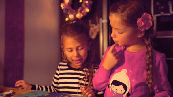 Zwei Mädchen sitzen am Tisch in einem weihnachtlich geschmückten Raum und lachen fröhlich.