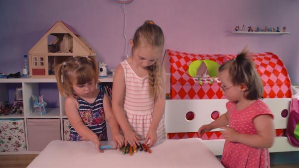 Drei Mädchen stehen in einem Kinderzimmer vor einem Tisch und wählen verschiedene Buntstifte aus.