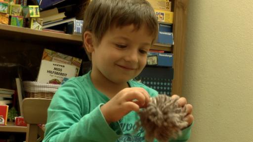 Ein Kind sitzt vor einem Regal und in der Hand hält es einen braunen Wollpompom.