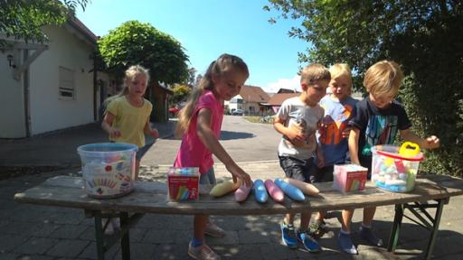 Fünf Kinder stehen draußen an einem Tisch und greifen nach der darauf liegenden Kreide.